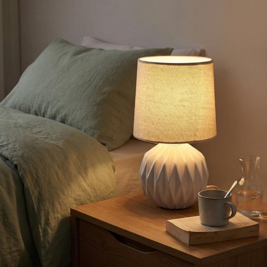 White geometric ceramic table lamp with linen shade casting warm ambient light on a bedroom nightstand.