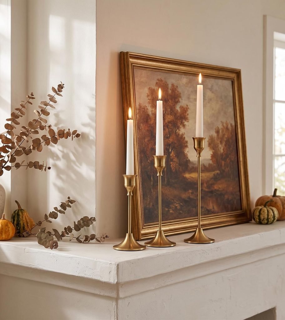 White fireplace mantel styled with staggered brass taper candlestick holders and framed autumn landscape art.
