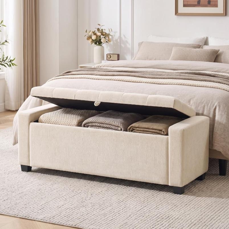 Beige tufted upholstered storage bench at the foot of a bed with open lid showing stored blankets.